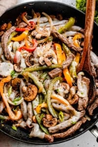 overhead shot of steak strips pepper strips, and mushrooms in a black skillet with brown wooden spoon mixing through the ingredients