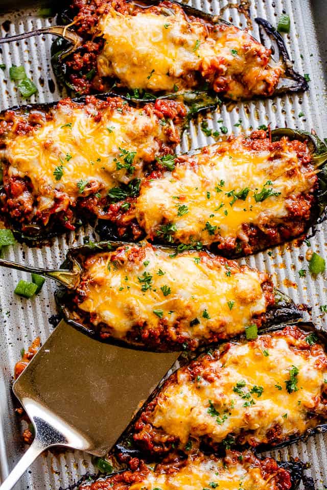 Overhead view of stuffed poblano peppers filled with chili and topped with melted cheese, with a spatula slid underneath one of the peppers.