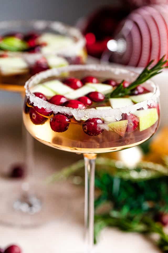 Close-up shot of two coupe glasses filled with prosecco and garnished with fresh cranberries, apples, and rosemary sprigs