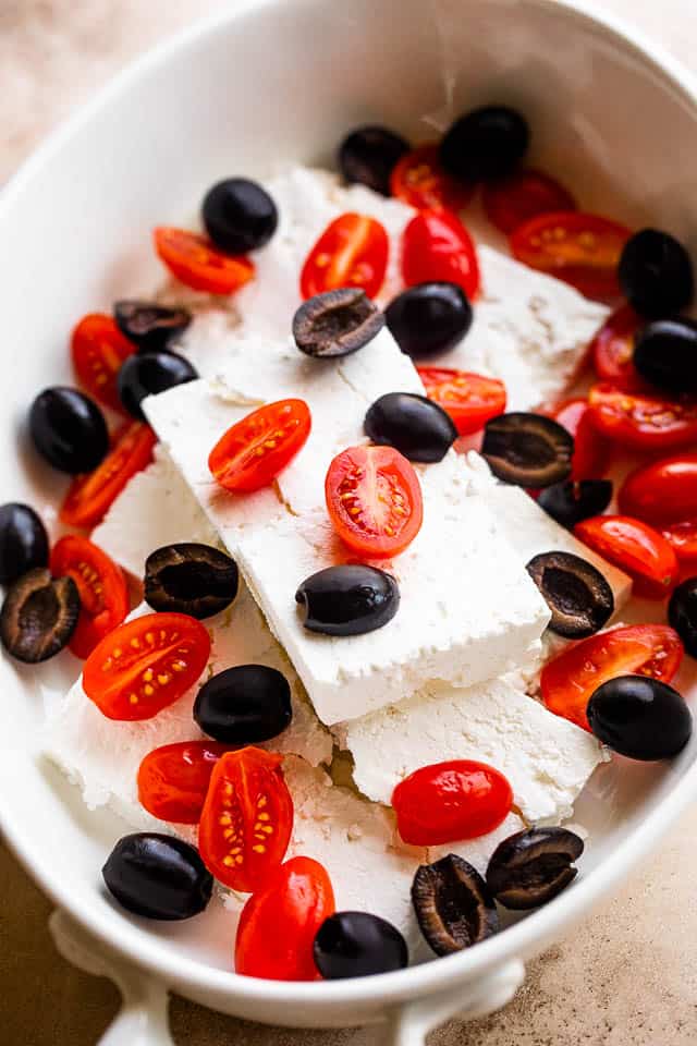 Blocks of feta in an oval baking dish and topped with olives and tomatoes.