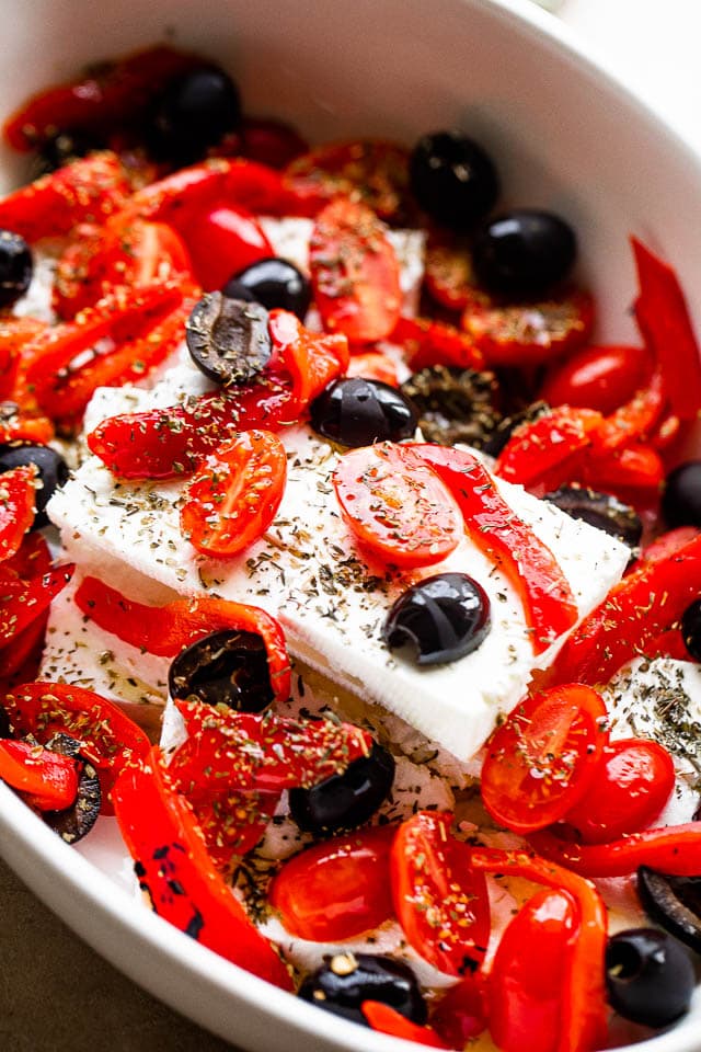 Blocks of feta in an oval baking dish and topped with olives and tomatoes.