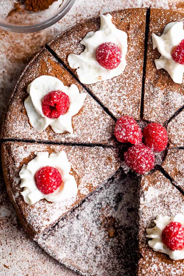 overhead close up shot of a sliced flourless chocolate cake topped with powdered sugar, berries, and cream