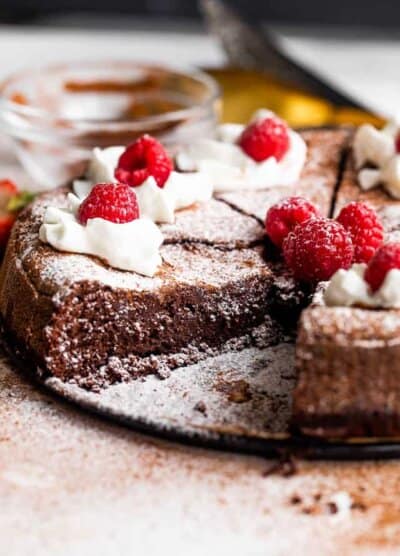 side shot of flourless chocolate cake cut into slices and topped with powdered sugar, whipped cream, and raspberries