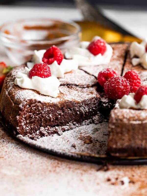 side shot of flourless chocolate cake cut into slices and topped with powdered sugar, whipped cream, and raspberries