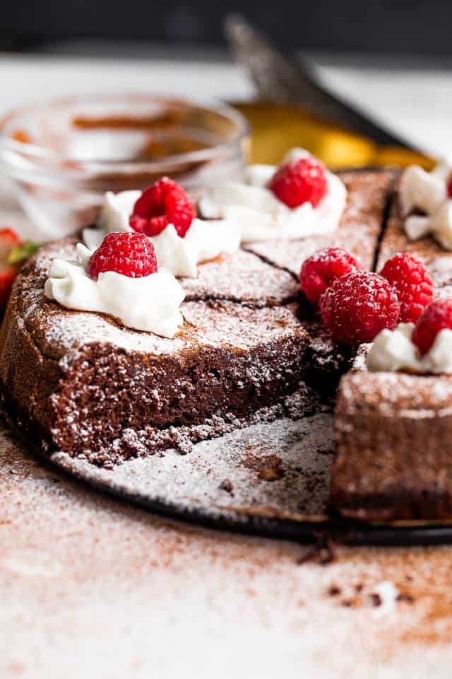 side shot of flourless chocolate cake cut into slices and topped with powdered sugar, whipped cream, and raspberries