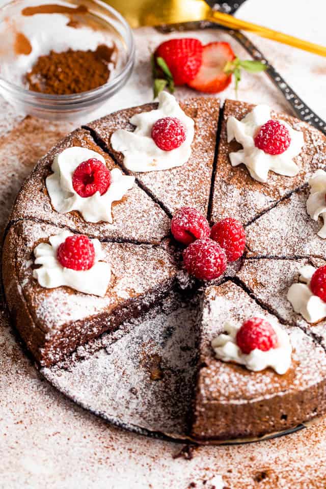 wide shot of a sliced flourless chocolate cake topped with powdered sugar, berries, and cream