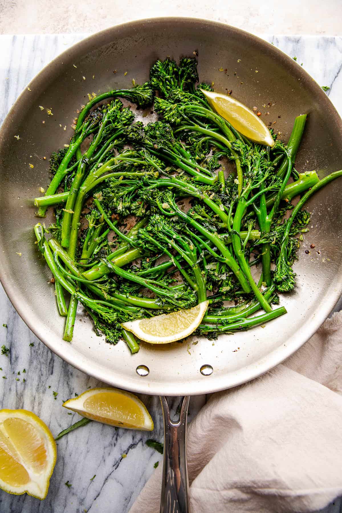 overhead shot of sauteed broccolini and lemon wedges pictured in a stainless steel skillet