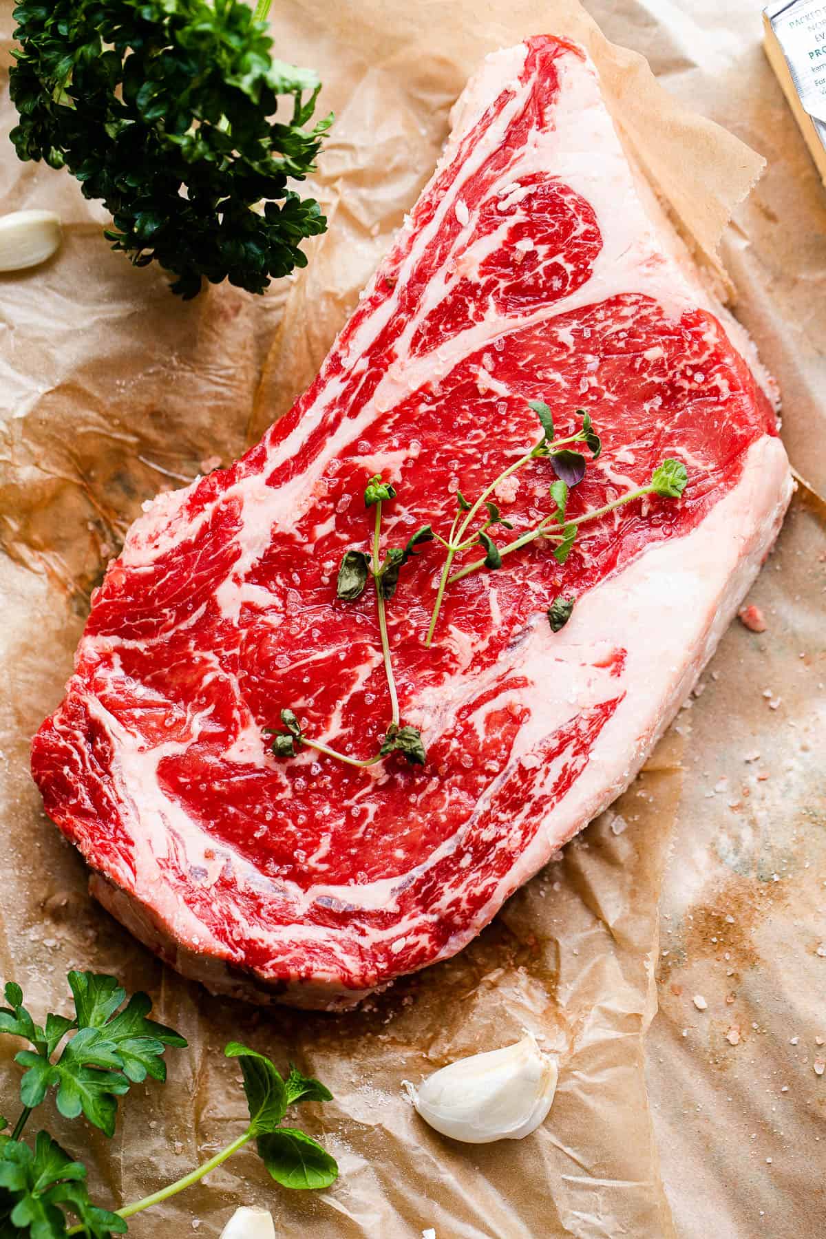 Raw ribeye steak set over brown paper with parsley and garlic cloves around it.