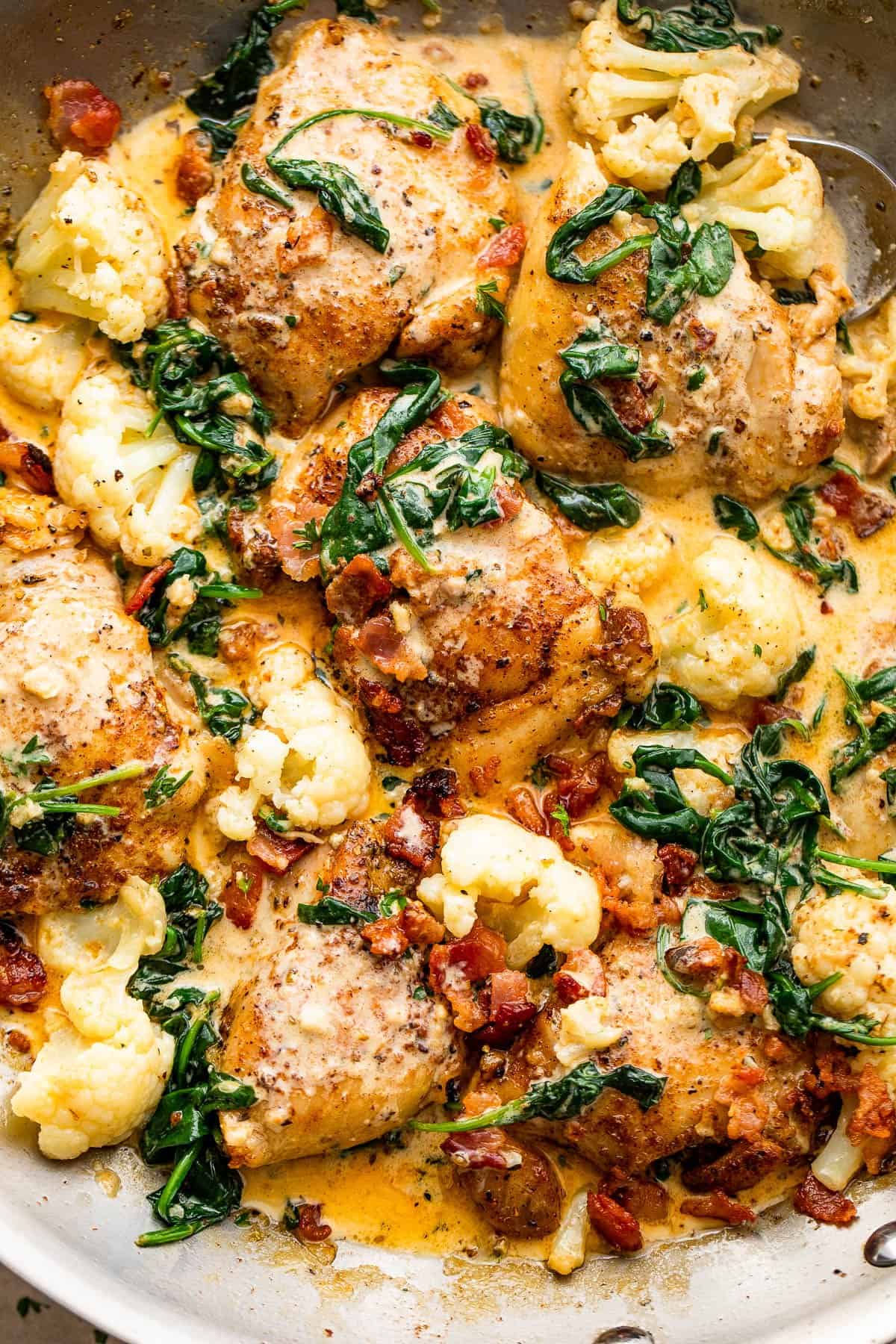 close up shot of cooked boneless chicken thighs topped with baby spinach, cream sauce, cauliflower, and bacon