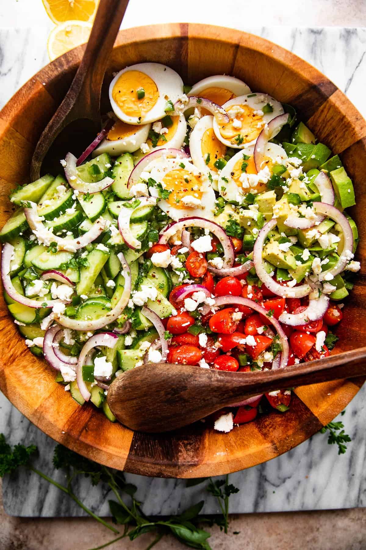 overhead shot of a wooden salad bowl filled with sliced red onions, halved hard boiled eggs, chopped avocado, sliced cherry tomatoes, and sliced cucumbers and topped with feta cheese