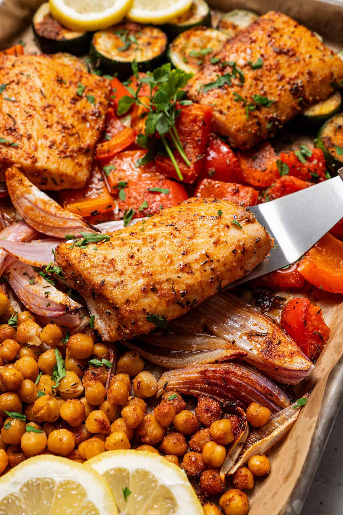 This easy Baked Mahi Mahi with Veggies is an all-in-one dinner that’s super easy to make! You’ll love the firm white fish, tender veggies and fragrant Mediterranean spices. Close up of a spatula lifting a baked mahi mahi filet from a sheet pan surrounded by chickpeas and roasted vegetables.