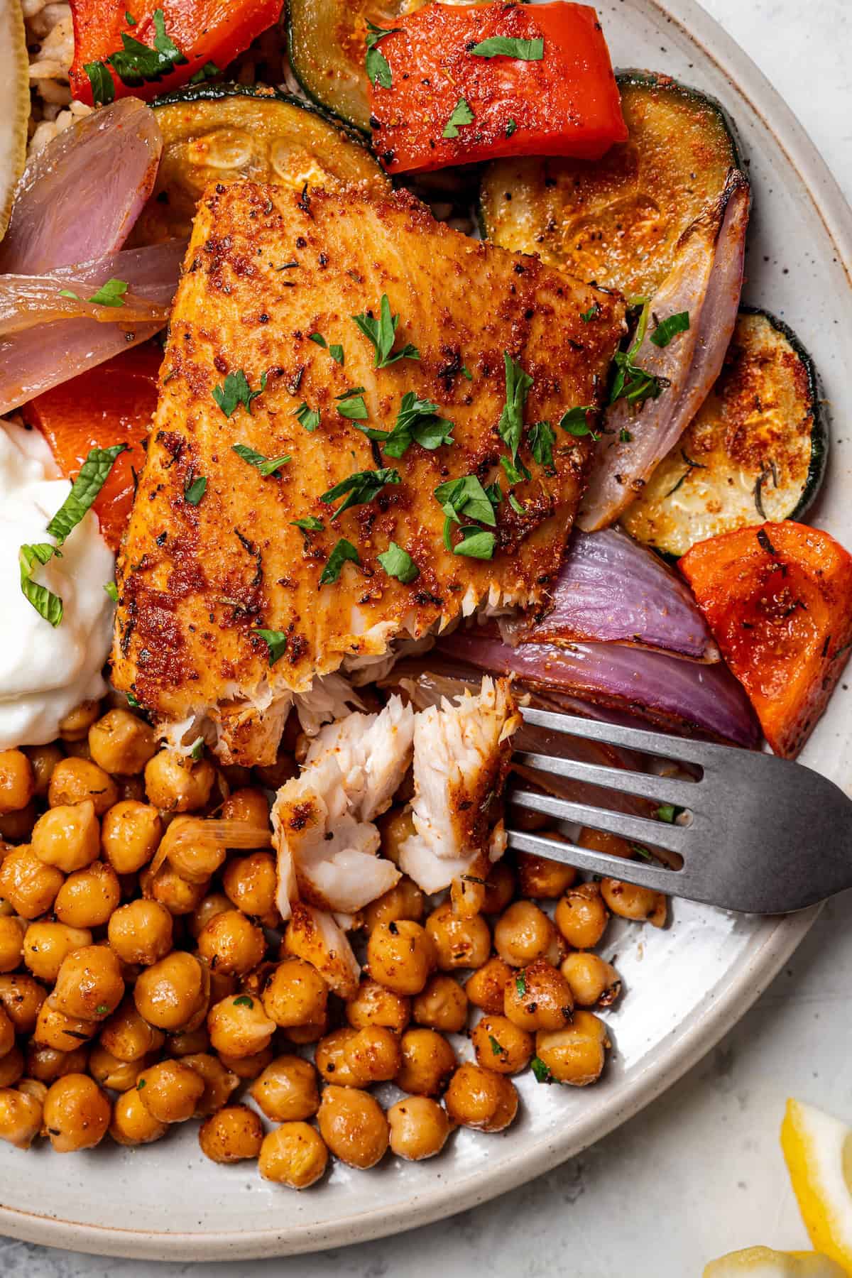 Overhead view of a fork flaking a piece of mahi mahi next to roasted chickpeas and Mediterranean veggies on a plate.