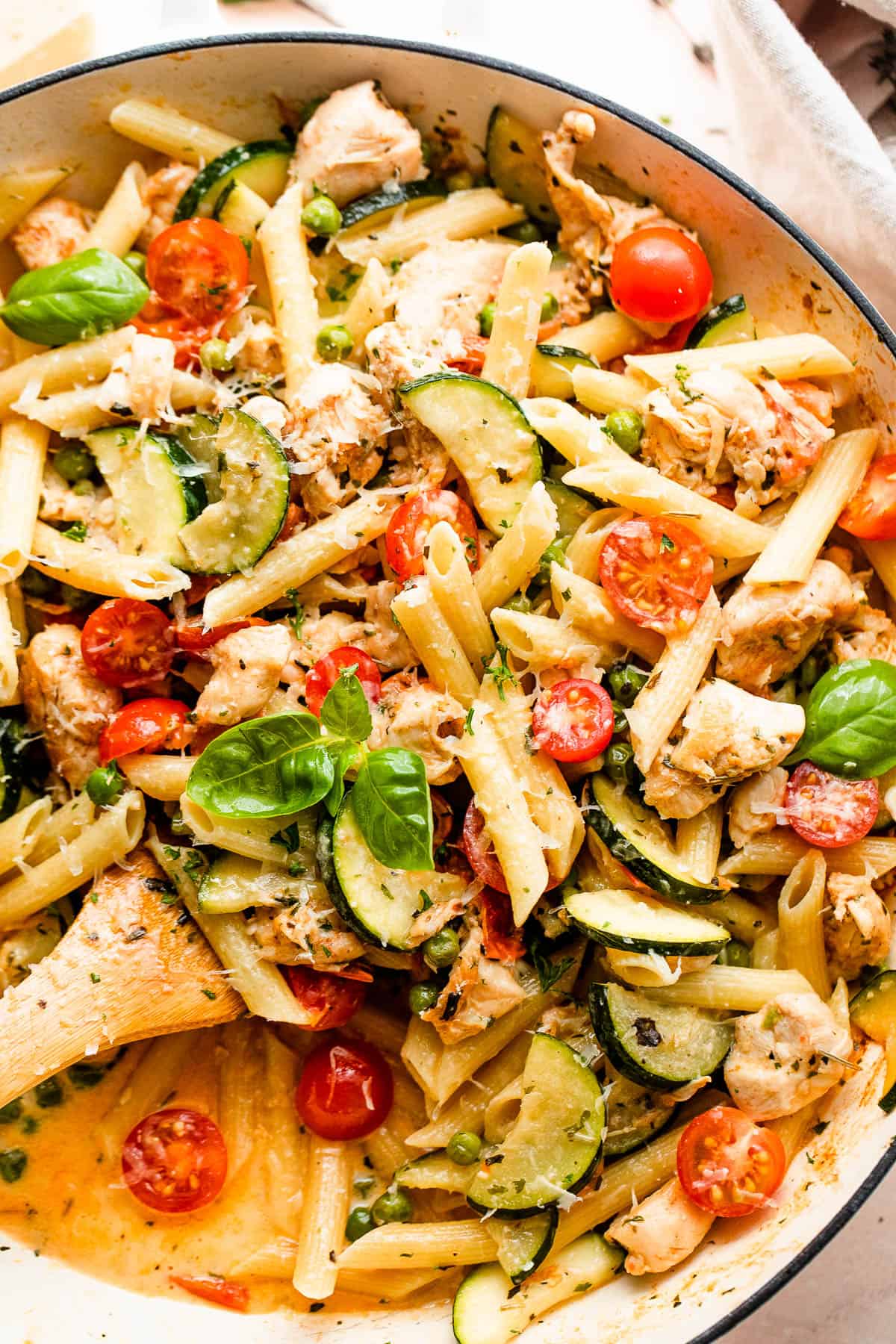 mixing penne pasta with zucchini, tomatoes, chicken, and peas in a skillet