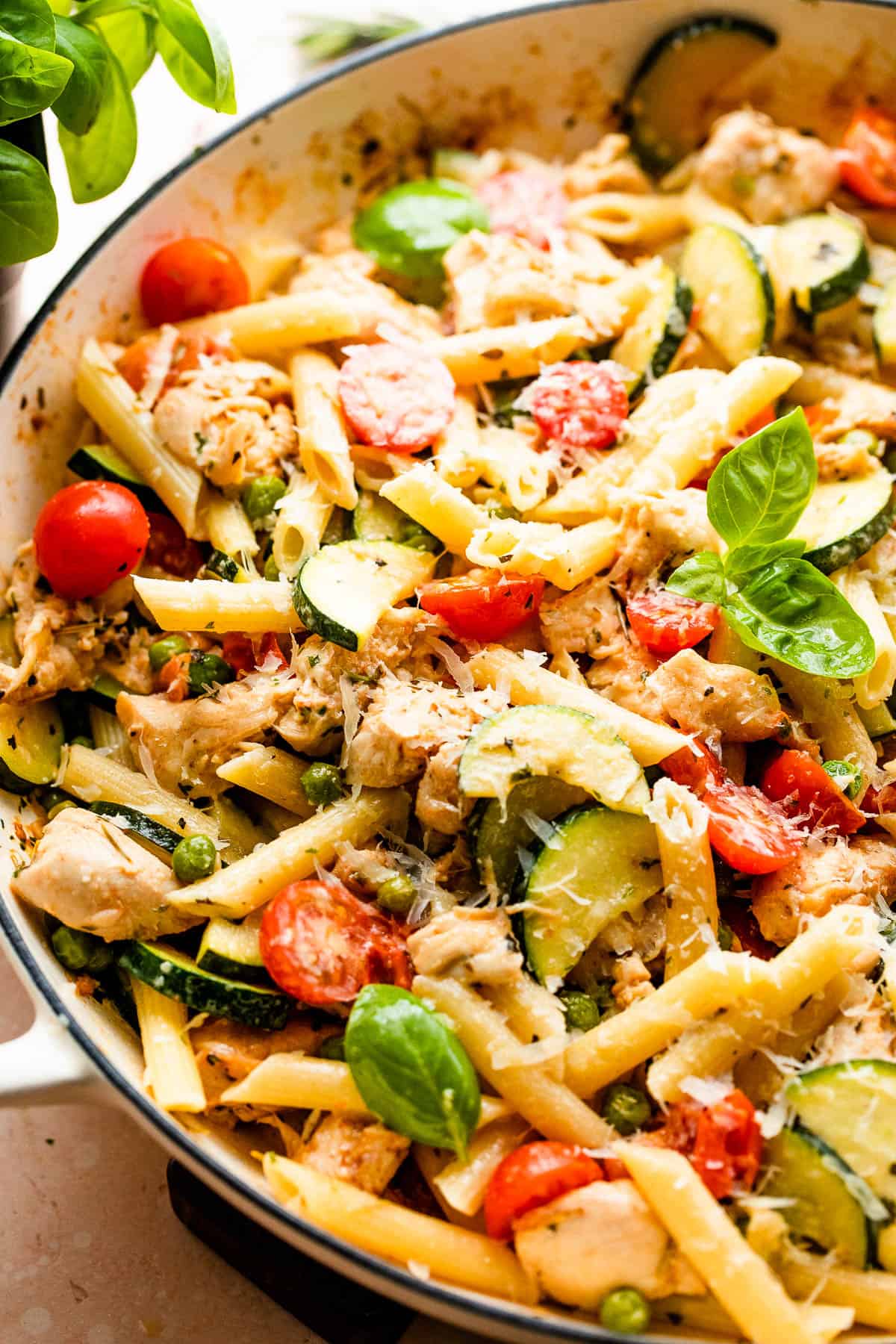 Close-up shot of penne pasta with zucchini, tomatoes, chicken, and peas in a skillet.