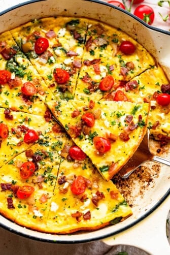 overhead shot of a sliced frittata in a white skillet topped with bacon and tomatoes