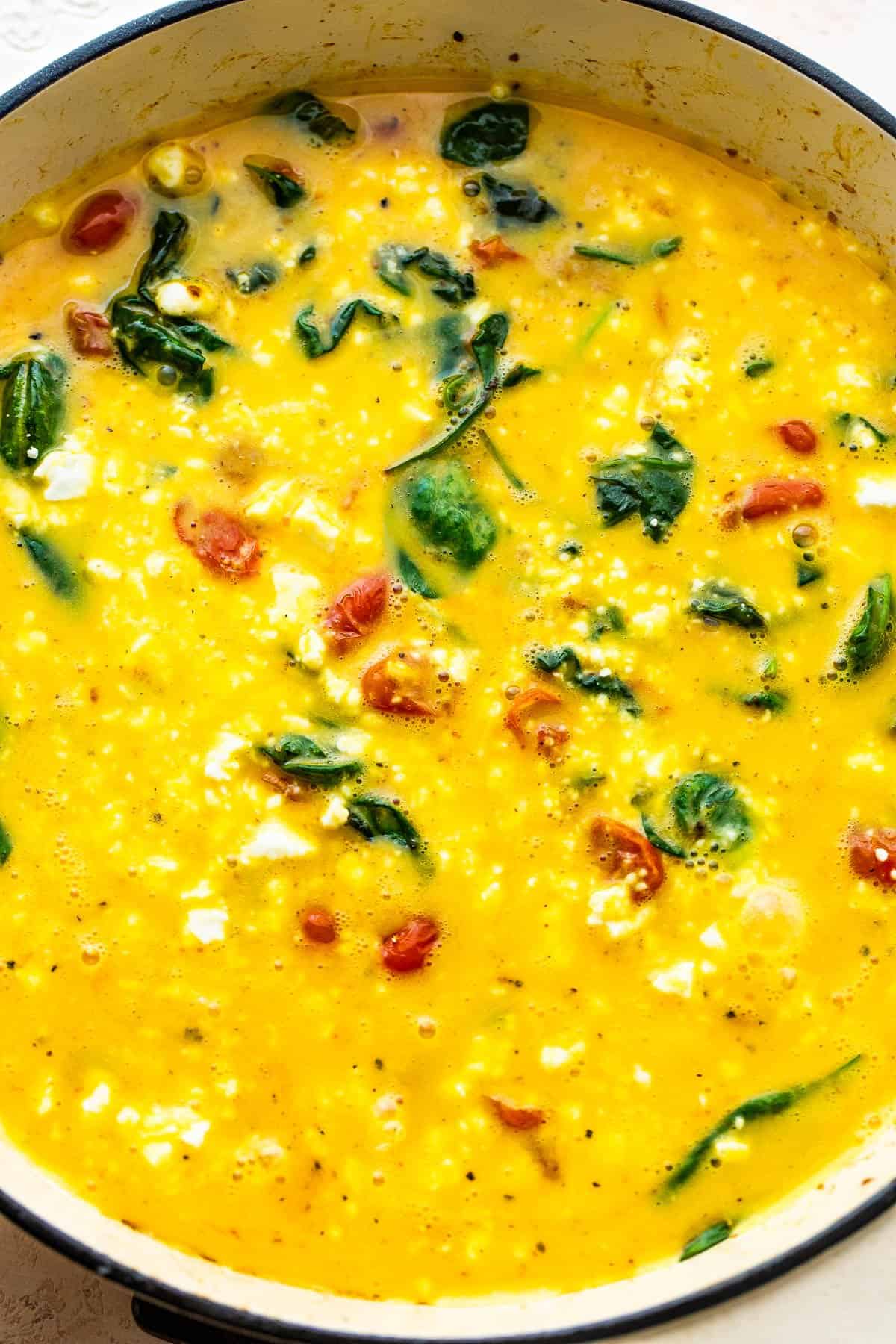 raw egg mixture with spinach and tomatoes poured into a white frying pan