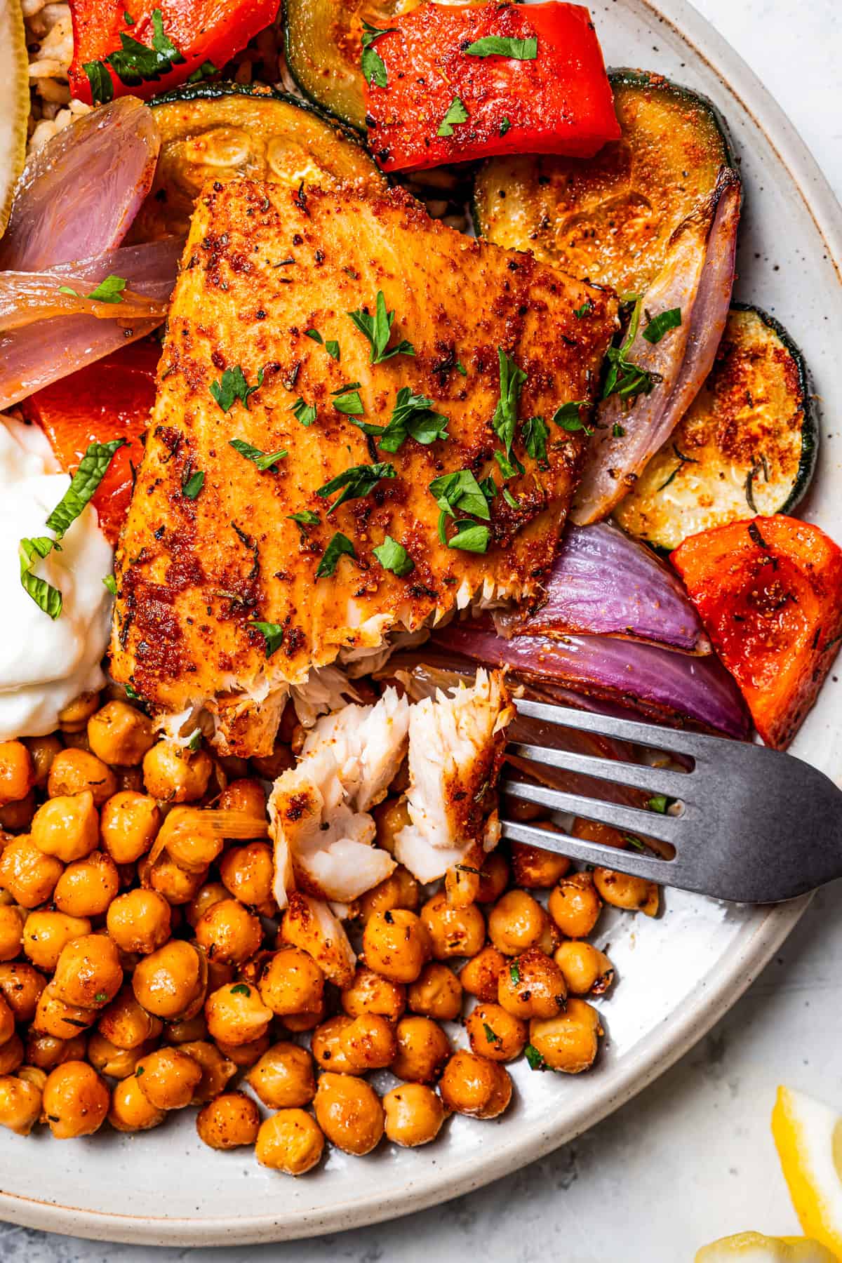 A fork flaking a piece of mahi mahi next to roasted chickpeas and veggies on a plate.