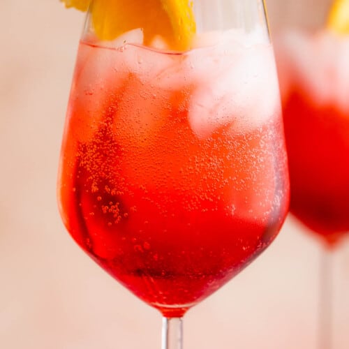 close up shot of Campari Spritz in a tall stemmed glass served with a slice of orange on the rim and red and white straw