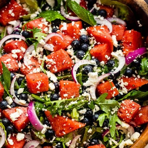 wooden salad bowl with cubed watermelon, blueberries, and sliced red onions atop a bed of torn up lettuce greens and topped with feta cheese crumbles