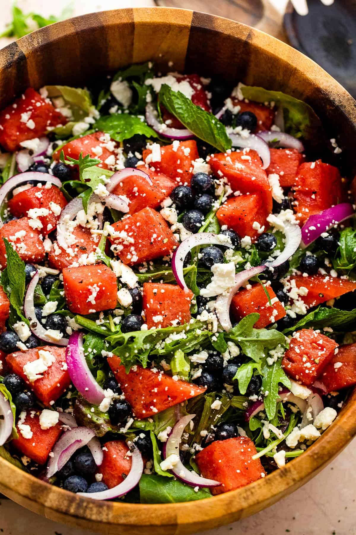 wooden salad bowl with cubed watermelon, blueberries, and sliced red onions atop a bed of torn up lettuce greens and topped with feta cheese crumbles