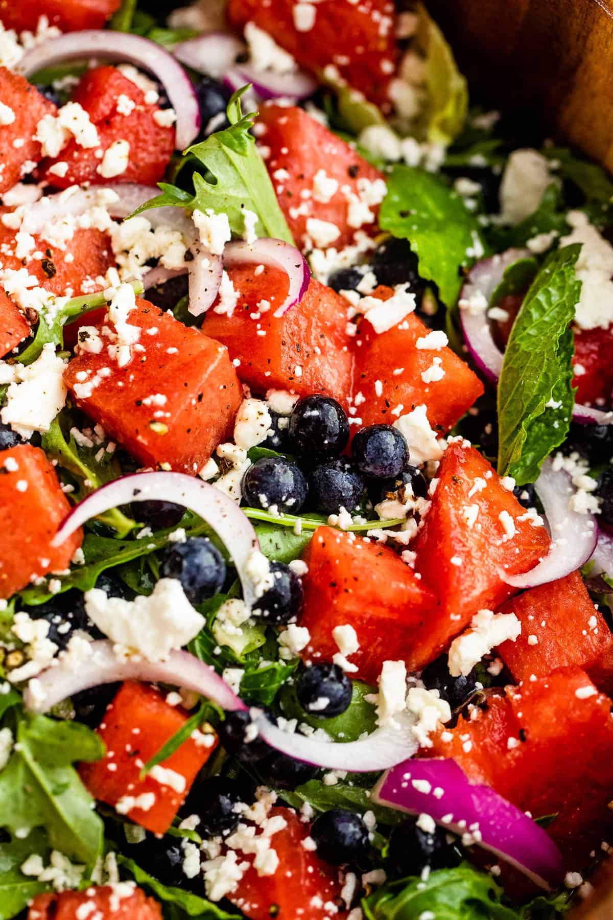 up close photo of cubed watermelon, blueberries, and sliced red onions atop a bed of torn up lettuce greens and topped with feta cheese crumbles