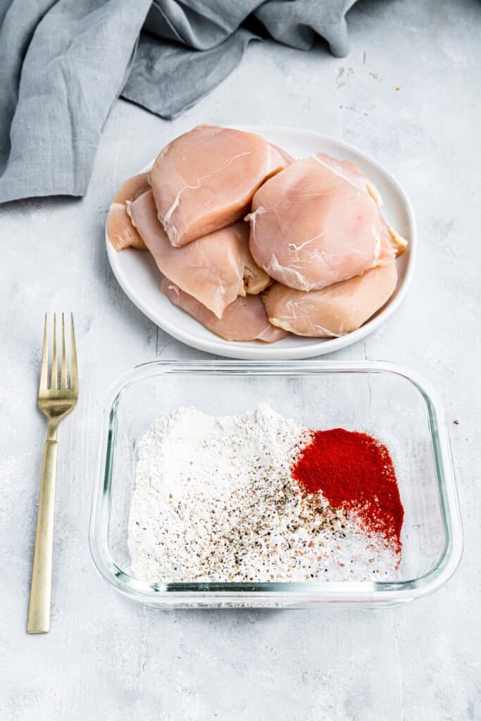 A Glass Bowl Filled with Flour, Paprika, Salt and Pepper Beside a White Plate Piled with Raw Chicken Breasts