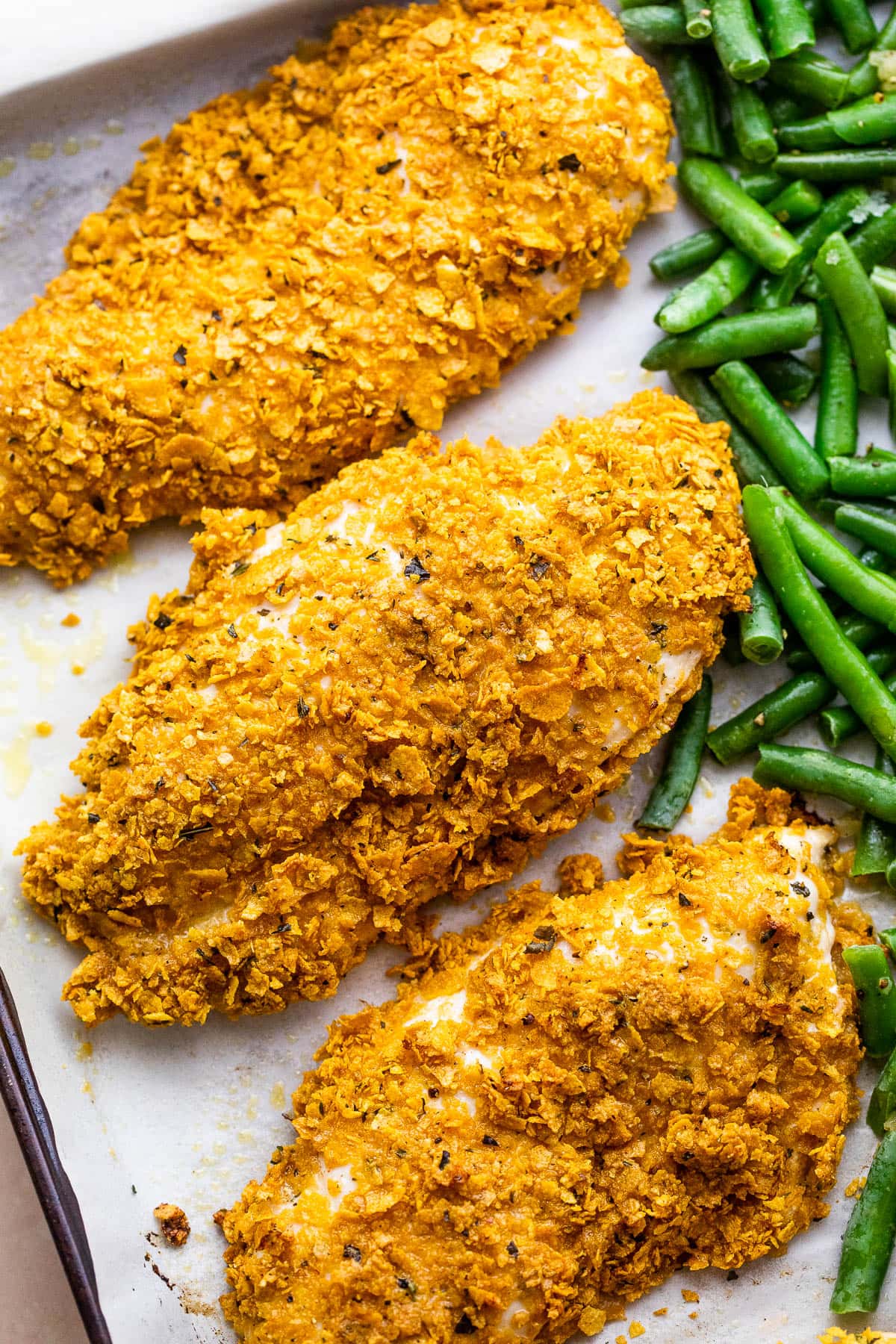 Baking sheet with three breaded chicken breasts and green beans arranged on the side of the chicken.