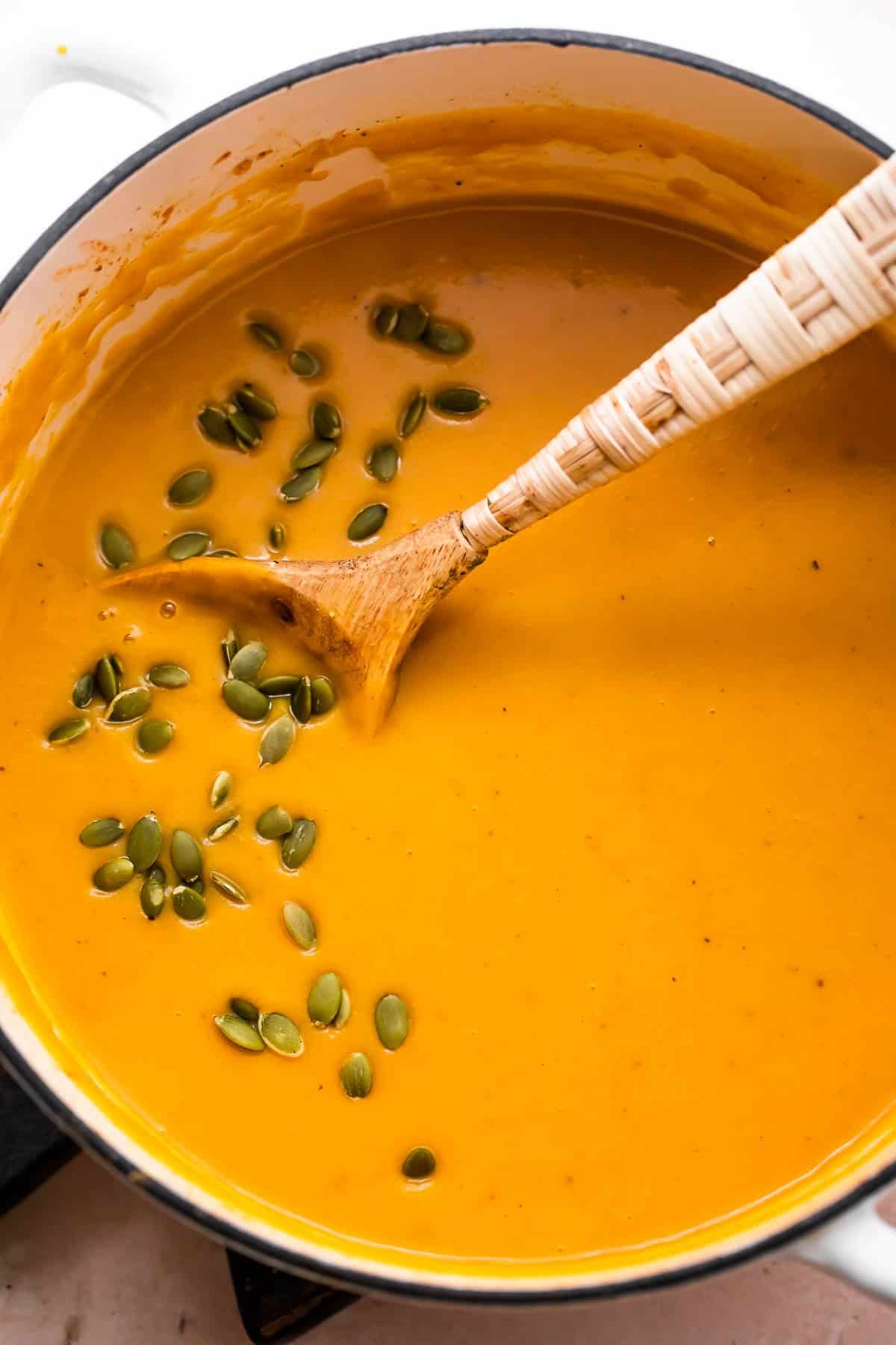 overhead shot of Curried Butternut Squash Soup in a Dutch oven with a wooden spoon inside the pot, and topped with a garnish ove pumpkin seeds