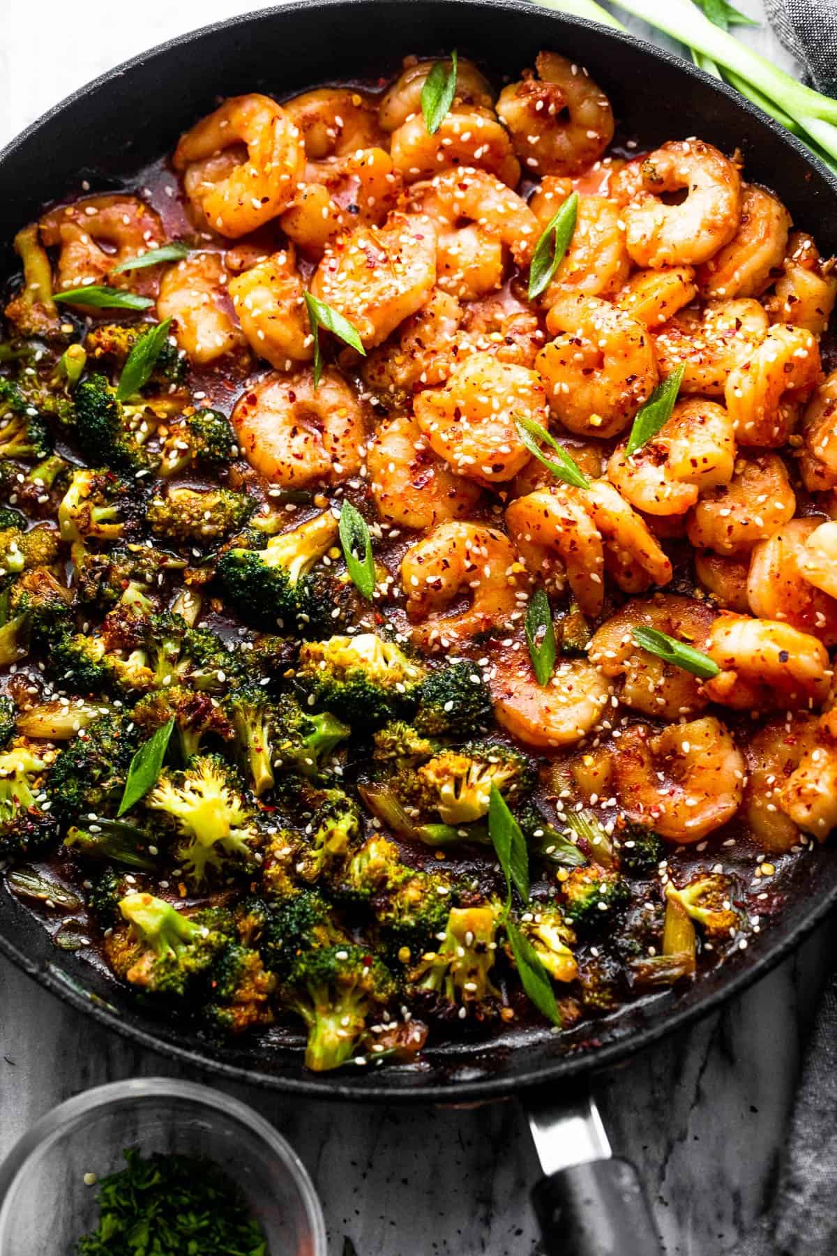 A large black skillet is full of broccoli on one side and shrimp on the other side, all sprinkled with sesame seeds and green onions.