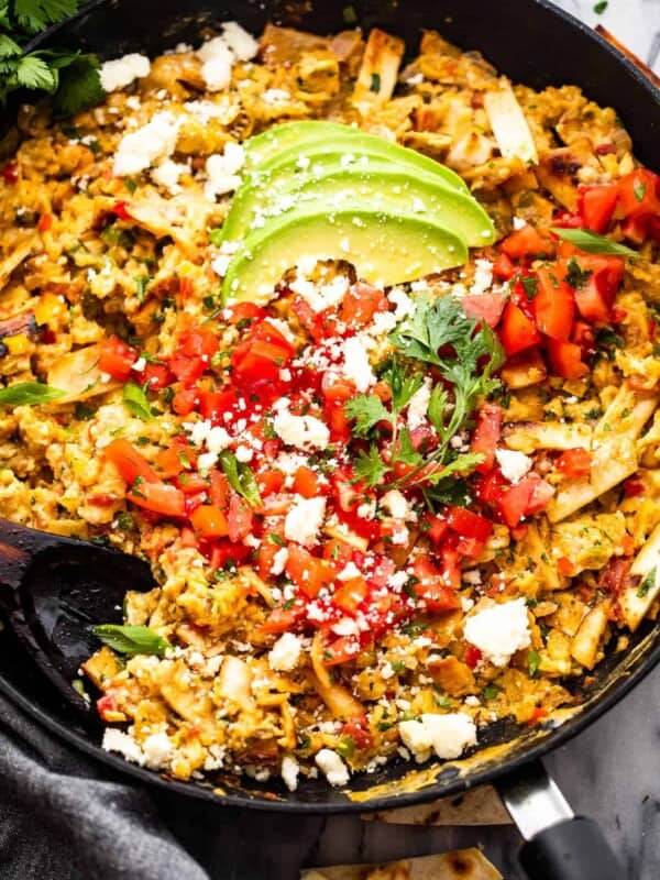 Migas in a skillet topped with sliced avocados, diced tomatoes, and cotija cheese