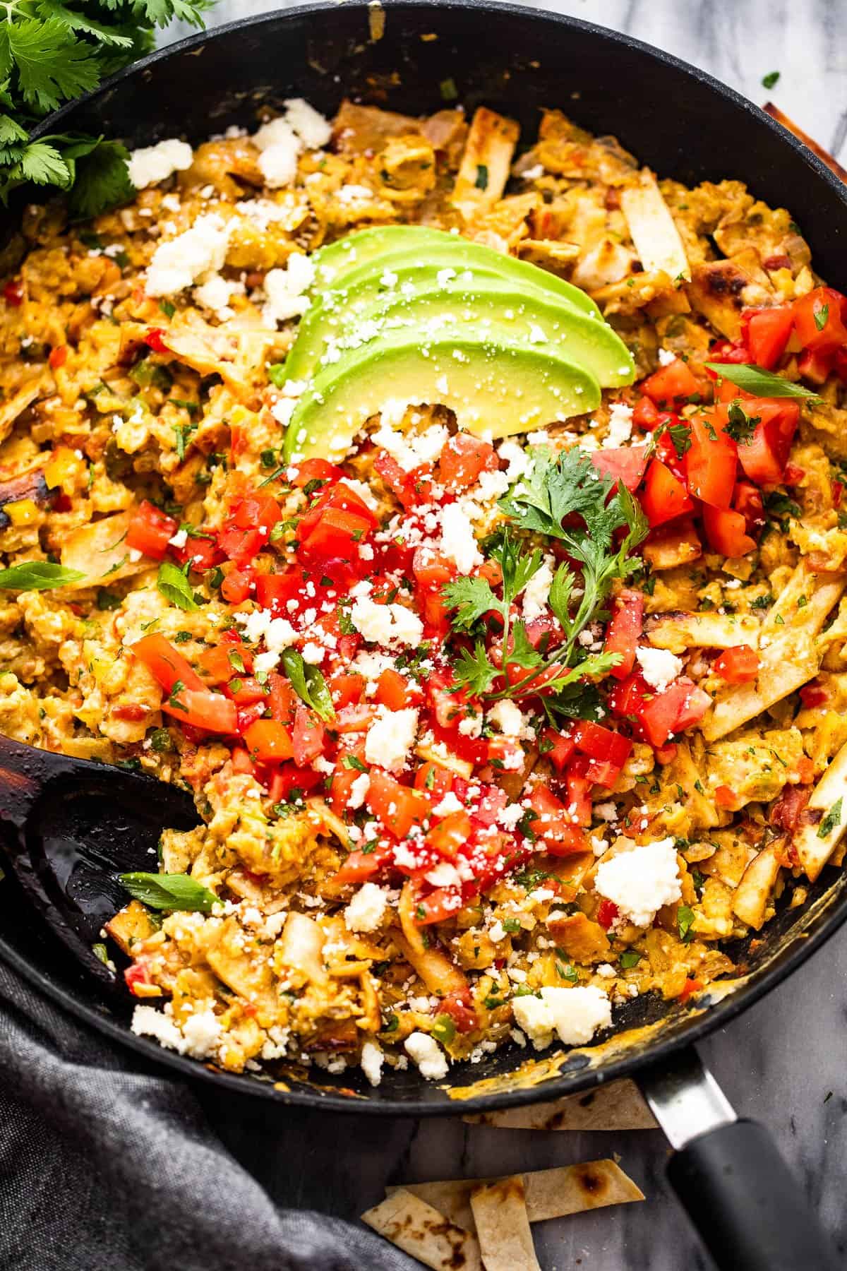 Migas in a skillet topped with sliced avocados, diced tomatoes, and cotija cheese