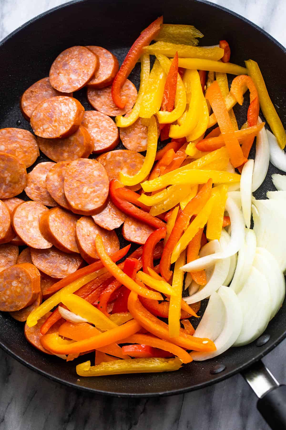 Sliced onions, peppers, and andouille sausages are placed in a black skillet before cooking them.