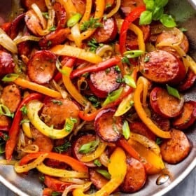 stainless steel skillet with cooked sliced peppers, sausages, and onions