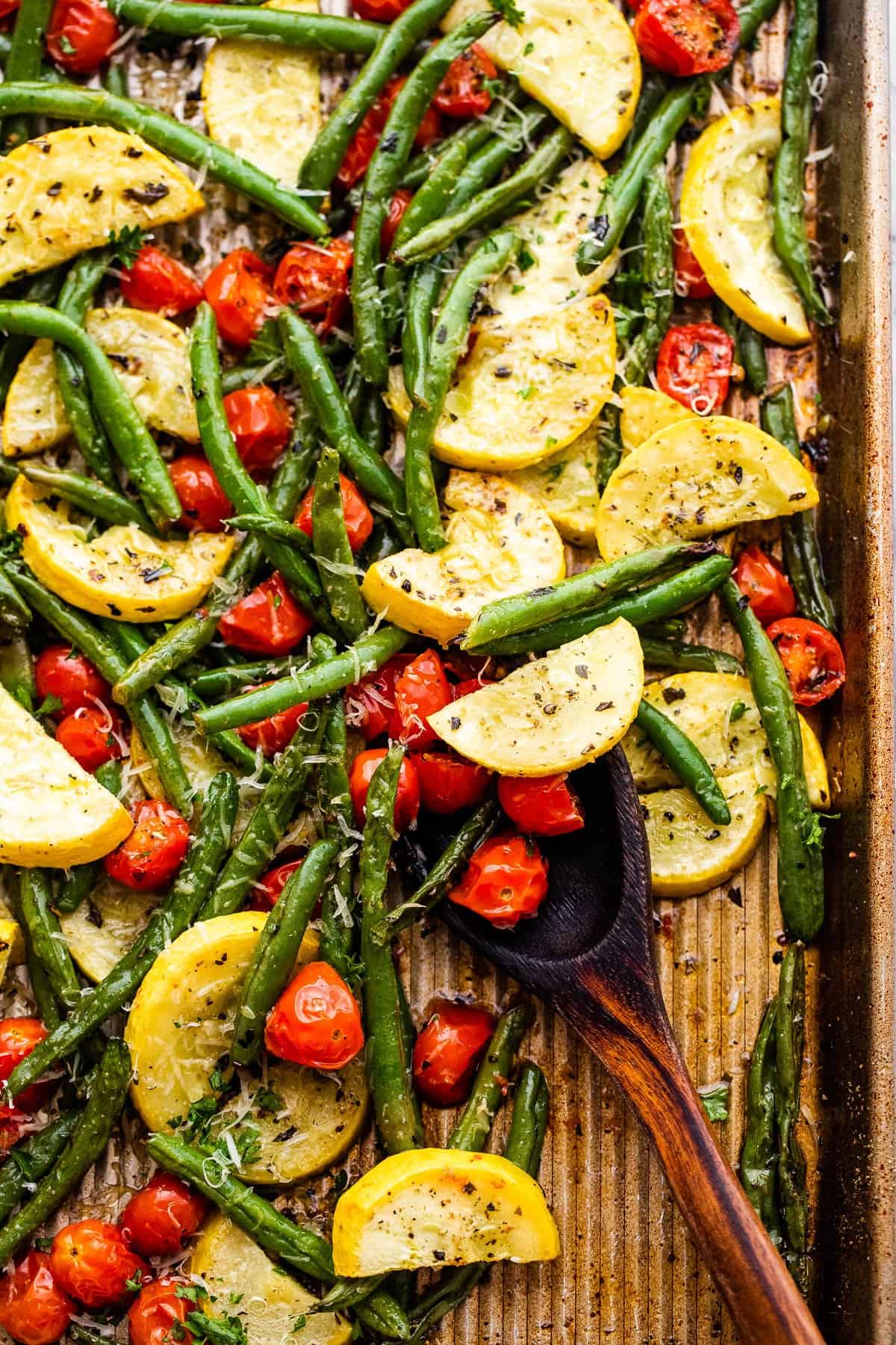 wooden spoon stirring through roasted cherry tomatoes, yellow squash, and green beans on a baking sheet.