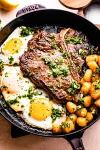 skillet with t-bone steak with over-easy eggs and tater tots arranged around it.