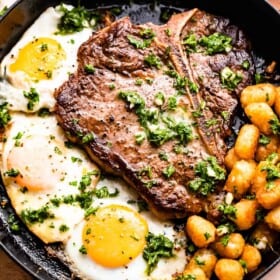 skillet with t-bone steak with over-easy eggs and tater tots arranged around it.