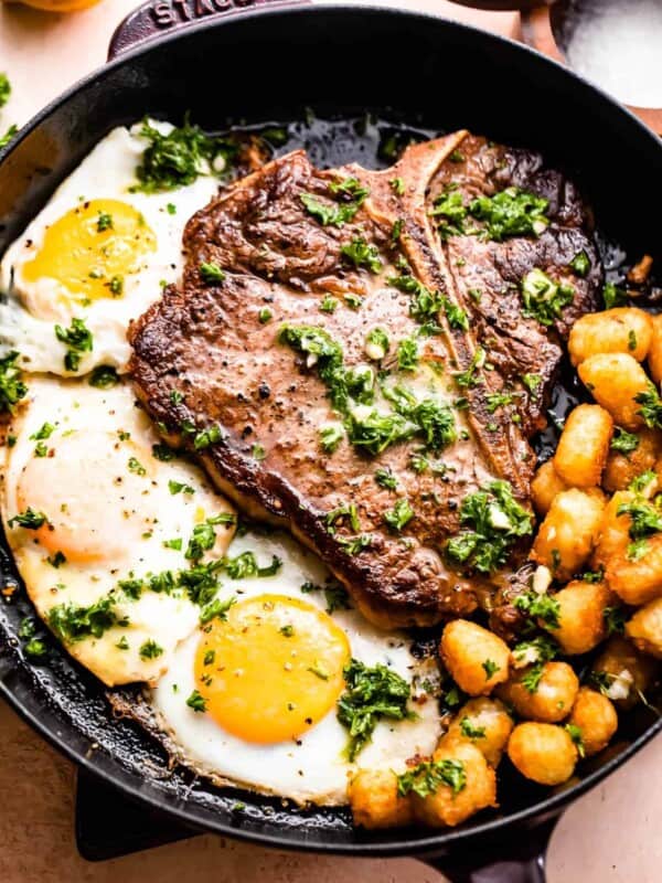 skillet with t-bone steak with over-easy eggs and tater tots arranged around it.
