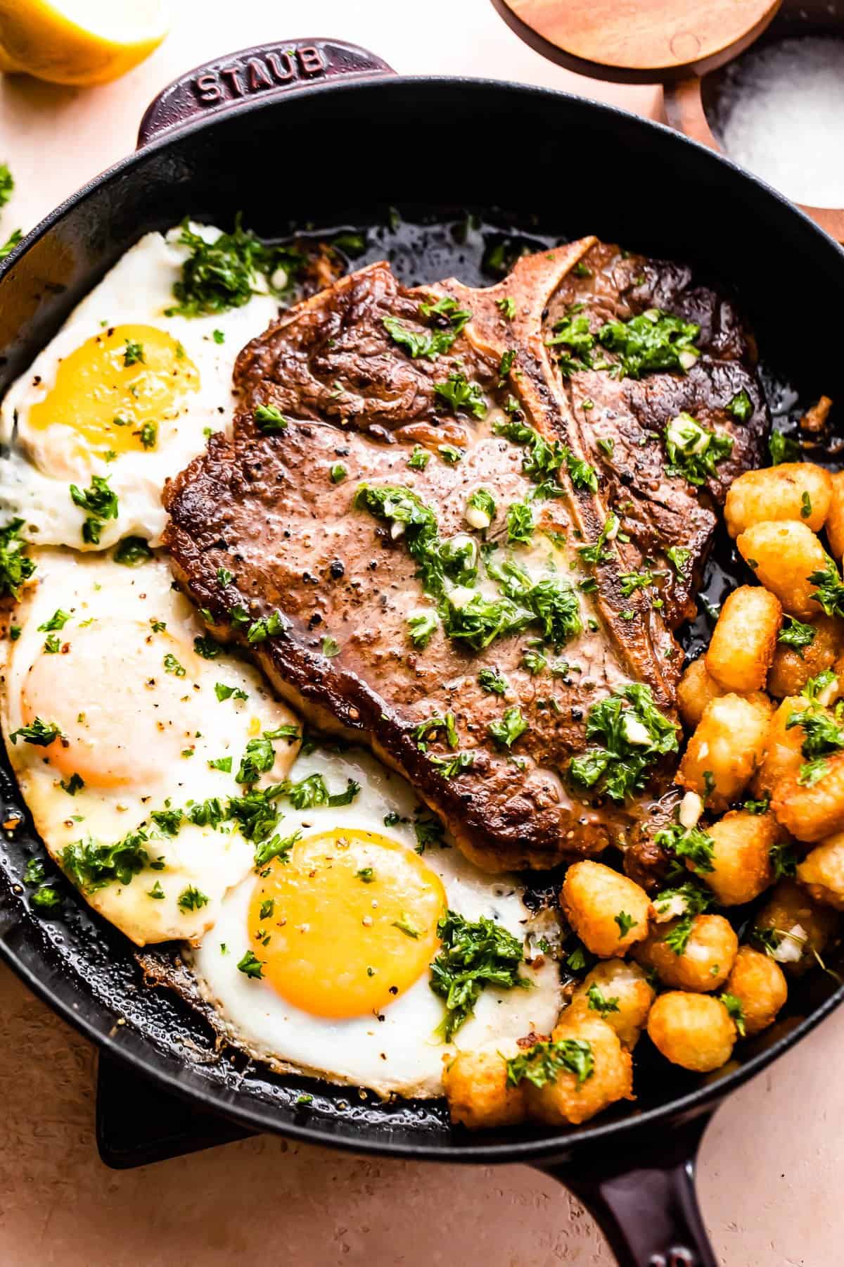 Skillet with t-bone steak with over-easy eggs and tater tots arranged around it.