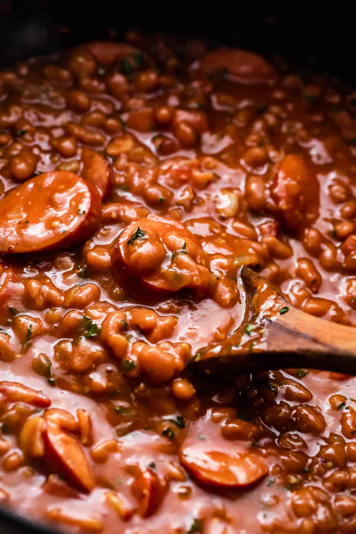 side shot of wooden spoon stirring through baked beans and sausage.
