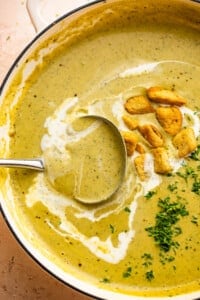 ladle inside a dutch oven with cream of zucchini soup, garnished with croutons, heavy cream swirl, and fresh green herbs