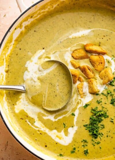 ladle inside a dutch oven with cream of zucchini soup, garnished with croutons, heavy cream swirl, and fresh green herbs