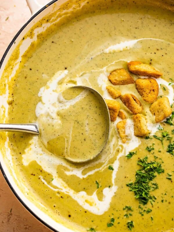 ladle inside a dutch oven with cream of zucchini soup, garnished with croutons, heavy cream swirl, and fresh green herbs