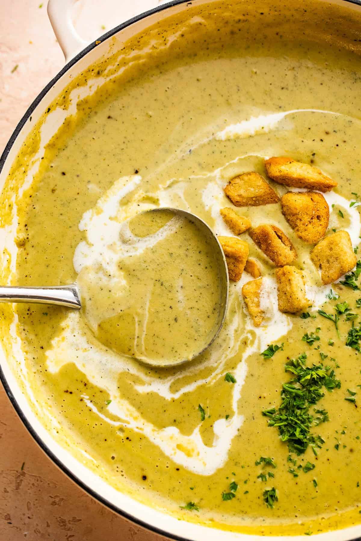 ladle inside a dutch oven with cream of zucchini soup, garnished with croutons, heavy cream swirl, and fresh green herbs