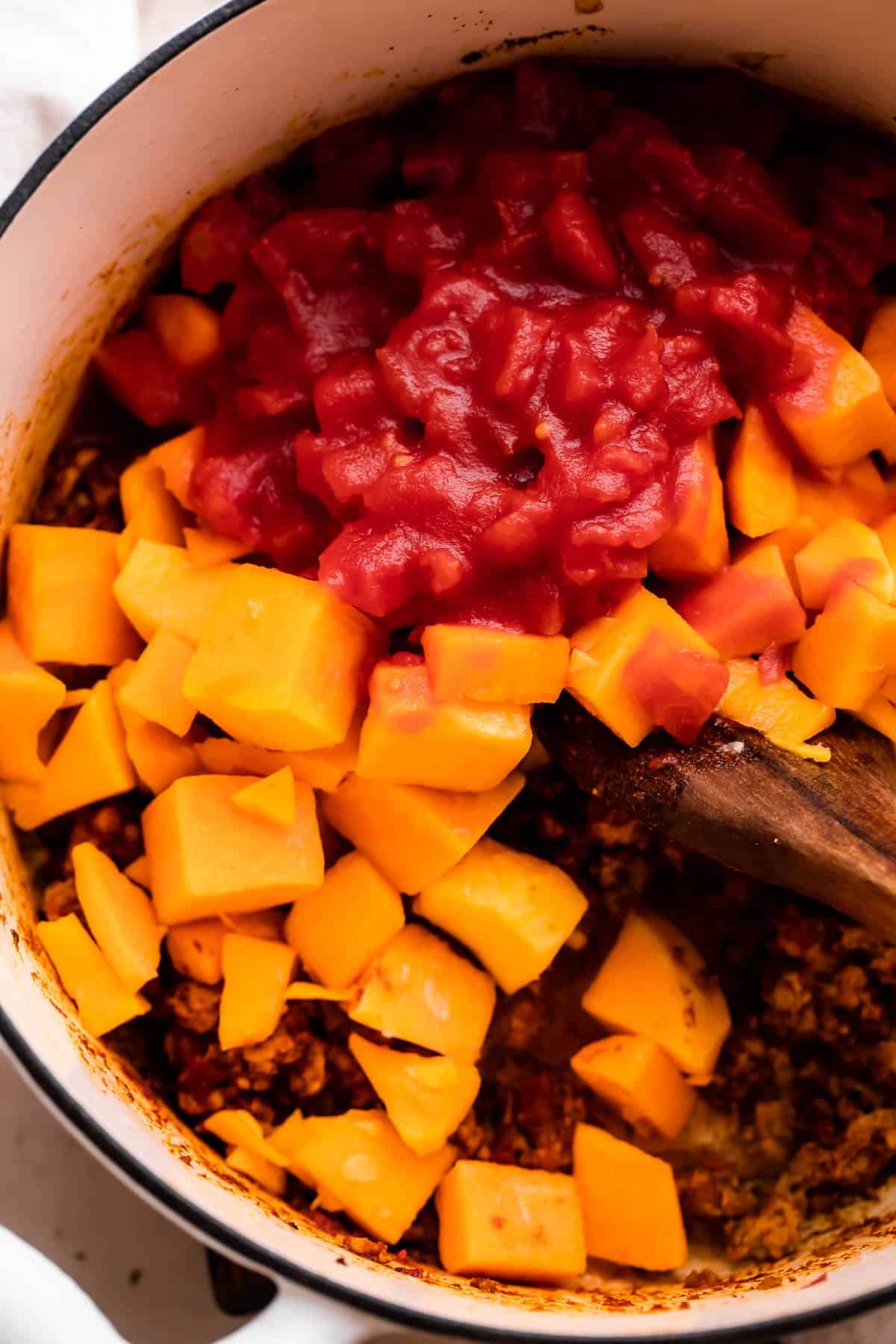 Stirring through cubed butternut squash and diced tomatoes in a dutch oven.