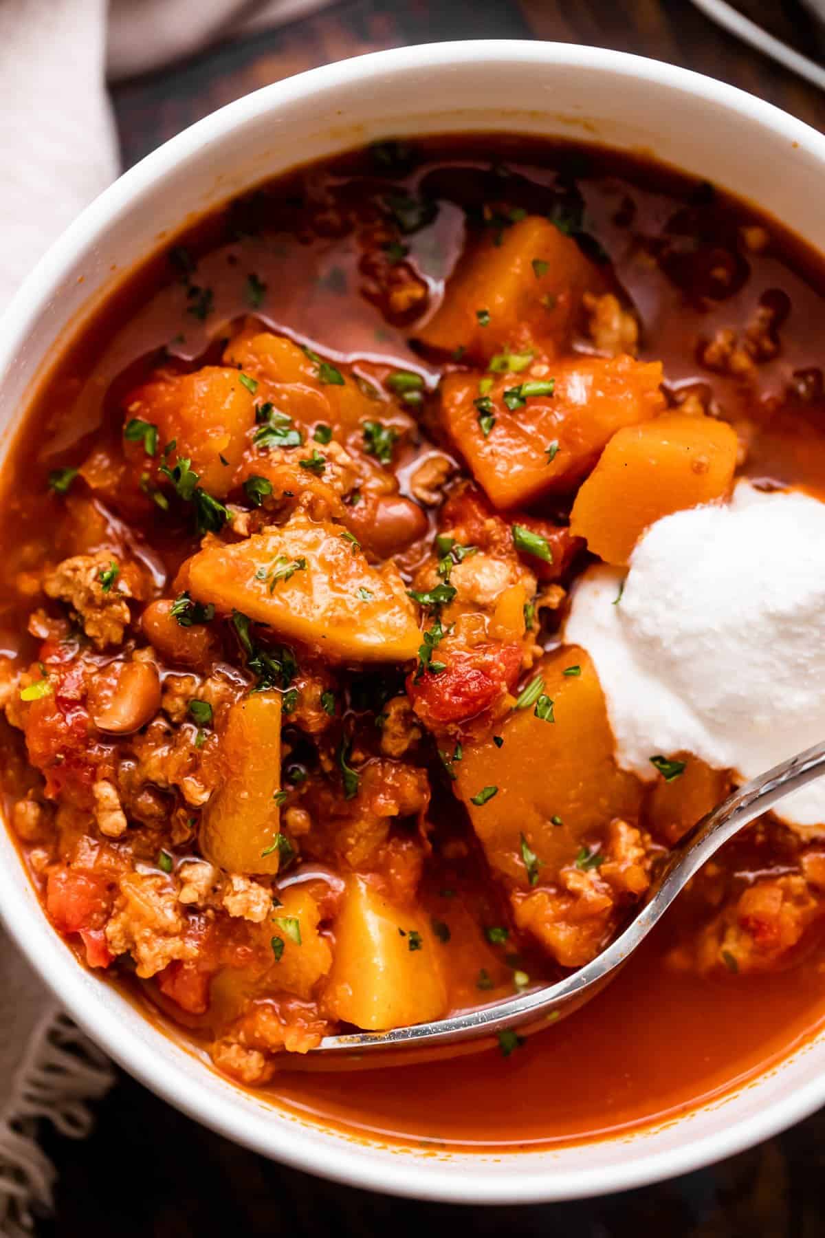 close up shot of a bowl with turkey chili, studded with cubed butternut squash, tomatoes, and a garnish of sour cream.