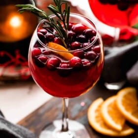 two tall stemmed glasses set on a wooden board and filled with wine, cranberries, orange slices, and rosemary sprigs.