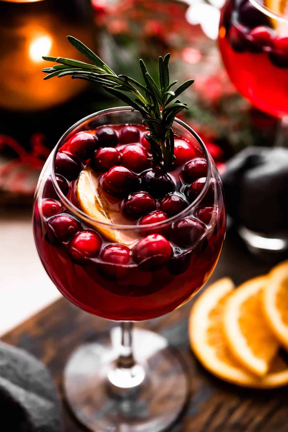 tall stemmed glass set on a wooden board and filled with wine, cranberries, orange slices, and rosemary sprigs.