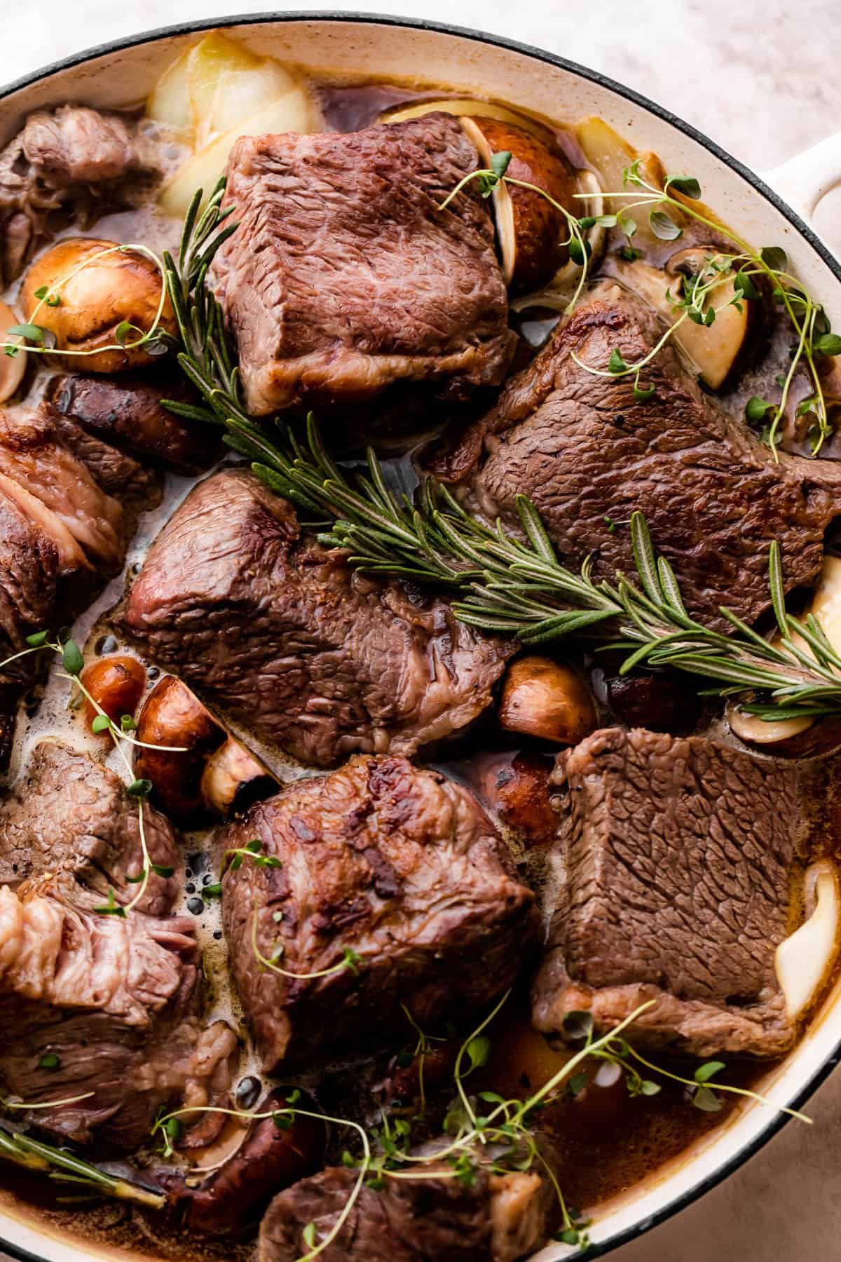 Browned beef chunks are placed in a white braiser with vegetables and rosemary sprigs.