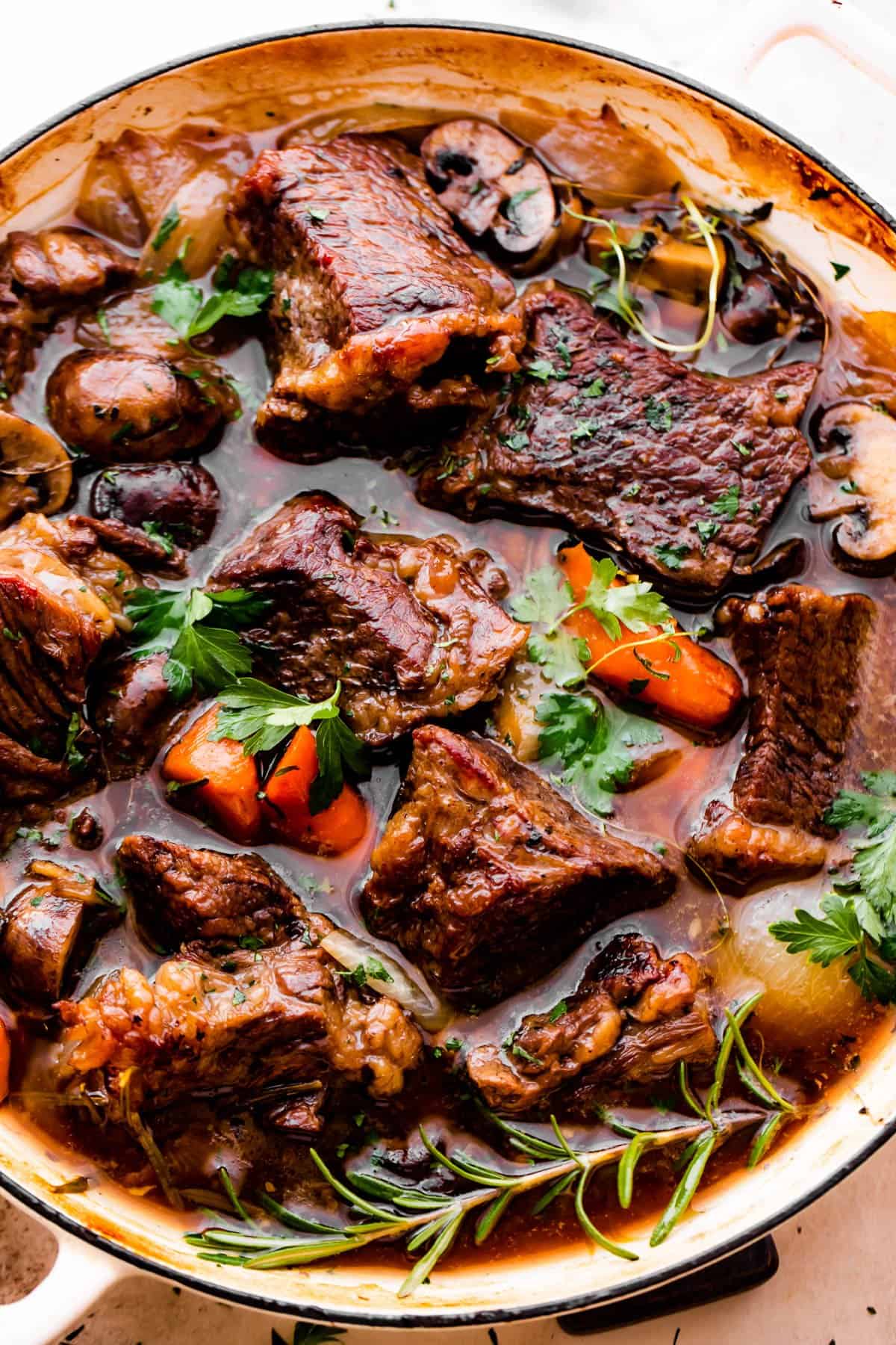 overhead shot of a white braiser filled with chunks of beef, carrots, mushrooms, and a garnish of parsley and rosemary sprigs.