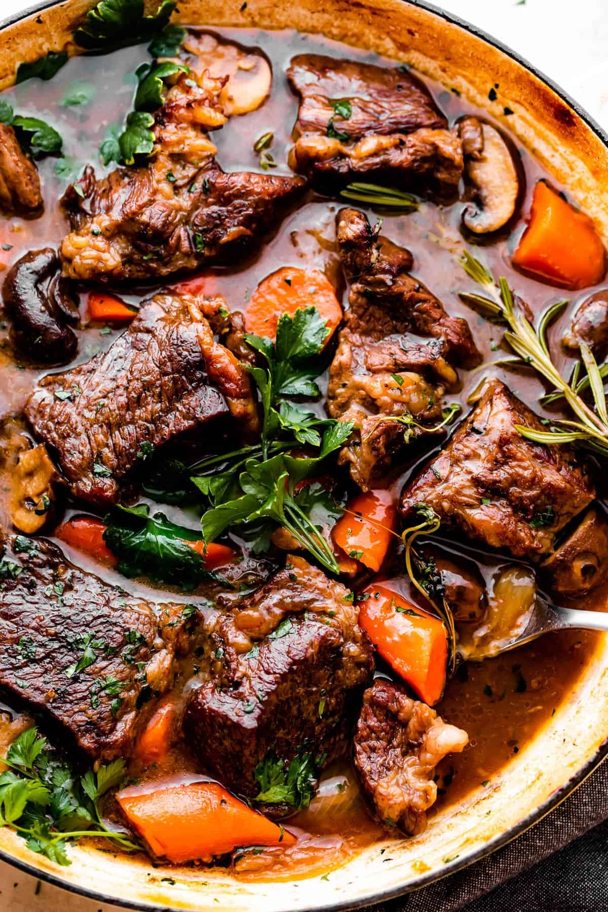 Overhead shot of Dutch oven pot roast in a braiser, filled with chunks of beef, carrots, mushrooms, and a garnish of parsley and rosemary sprigs.